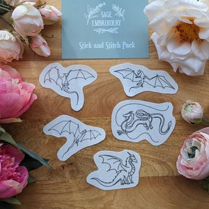 May include: Five white iron-on patches with black line drawings of dragons. The patches are arranged on a wooden surface. The text "Stick and Stitch Pack" is printed on a blue card in the background.