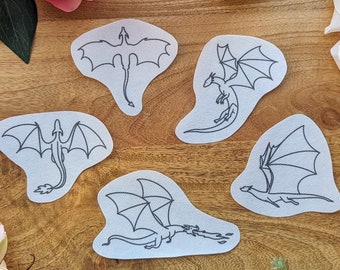 May include: Five black and white line drawings of dragons on white paper. The drawings are on a wooden surface with pink flowers around the edges.