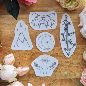 以下が含まれることがあります： 山、三日月、羽根のある人物、剣、星を持った手など、さまざまなデザインが施されたアイロン接着パッチ7個セット。パッチは白地に黒のアウトラインです。