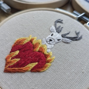 May include: Embroidered hoop art featuring a white deer head emerging from a heart-shaped flame. The flames are in shades of red, orange, and yellow. The deer has gray antlers. The embroidery is on a beige fabric.