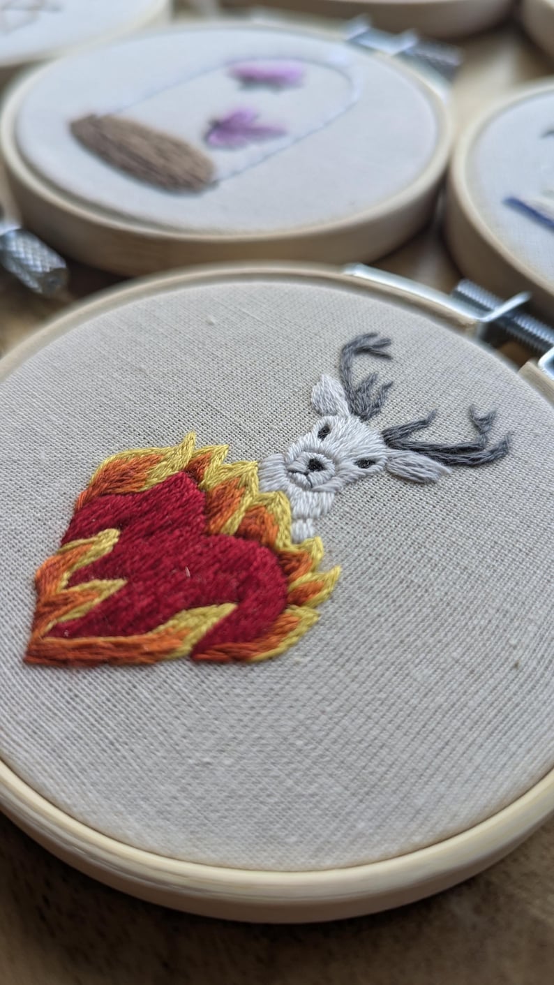 May include: Embroidered hoop art featuring a white deer head emerging from a heart-shaped flame. The flames are in shades of red, orange, and yellow. The deer has gray antlers. The embroidery is on a beige fabric.