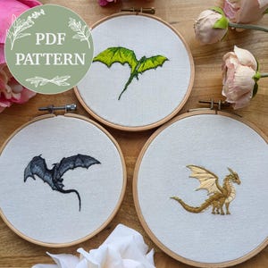 May include: Three embroidery hoops with dragon designs. One dragon is green, one is black, and the third is gold. The text "PDF PATTERN" is in a green circle in the upper left corner. The hoops are on a wooden surface with pink flowers.