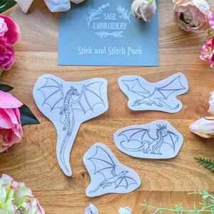 May include: A set of four white fabric dragon embroidery patterns with black outlines. The patterns are on a wooden surface with flowers around them. The card in the centre reads "Sage Embroidery Stick and Stitch Pack".
