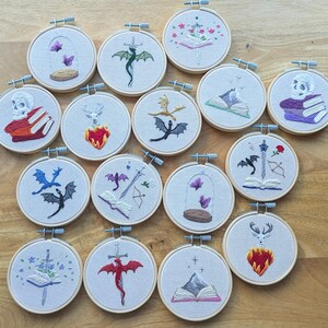 May include: A collection of embroidered hoops featuring various fantasy-themed designs. Each hoop showcases detailed needlework, including dragons, butterflies, skulls, and open books. The color palette includes shades of red, purple, green, and black, set against a neutral fabric background.