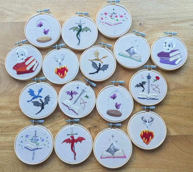 May include: A collection of embroidered hoops featuring various fantasy-themed designs. Each hoop showcases detailed needlework, including dragons, butterflies, skulls, and open books. The color palette includes shades of red, purple, green, and black, set against a neutral fabric background.