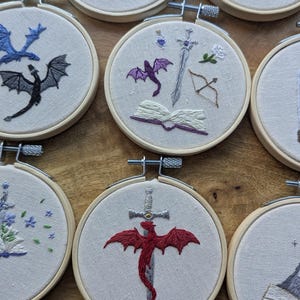May include: Embroidered hoop art featuring fantasy themes. Designs include dragons, swords, books, and other symbols, all stitched in vibrant colors. The hoops are set against a neutral fabric background, creating a cohesive collection.