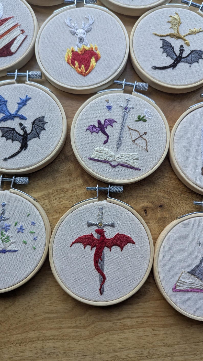 May include: Embroidered hoop art featuring fantasy themes. Designs include dragons, swords, books, and other symbols, all stitched in vibrant colors. The hoops are set against a neutral fabric background, creating a cohesive collection.