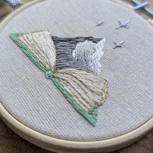 May include: Embroidered artwork in a wooden hoop. The design features a stylized mountain scene with a fan-shaped element in shades of beige, gray, and green. Small embroidered stars are scattered across the top. The fabric is a light beige.