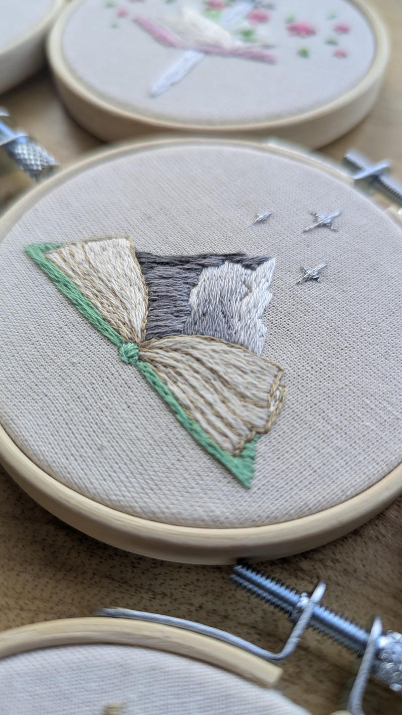 May include: Embroidered artwork in a wooden hoop. The design features a stylized mountain scene with a fan-shaped element in shades of beige, gray, and green. Small embroidered stars are scattered across the top. The fabric is a light beige.