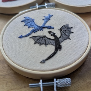 May include: Embroidered hoop art featuring two dragons, one blue and one black, on a beige fabric background. The dragons are detailed with wings and scales. The hoops are made of wood and are part of a collection.