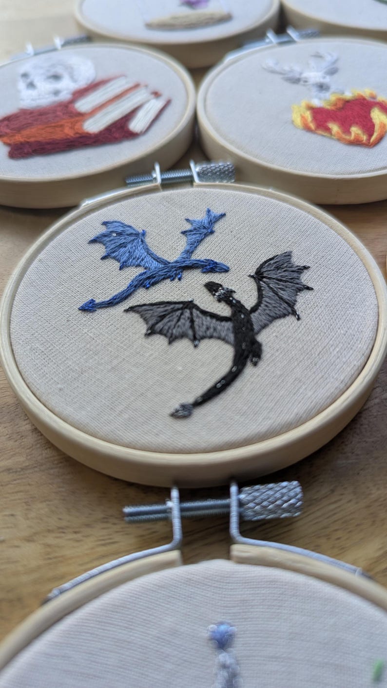 May include: Embroidered hoop art featuring two dragons, one blue and one black, on a beige fabric background. The dragons are detailed with wings and scales. The hoops are made of wood and are part of a collection.