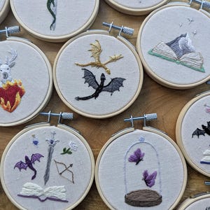 May include: A collection of embroidered hoops featuring fantasy-themed designs. Dragons, swords, books, and butterflies are depicted in colorful thread on a neutral fabric. Each hoop is secured with a metal tightening mechanism.