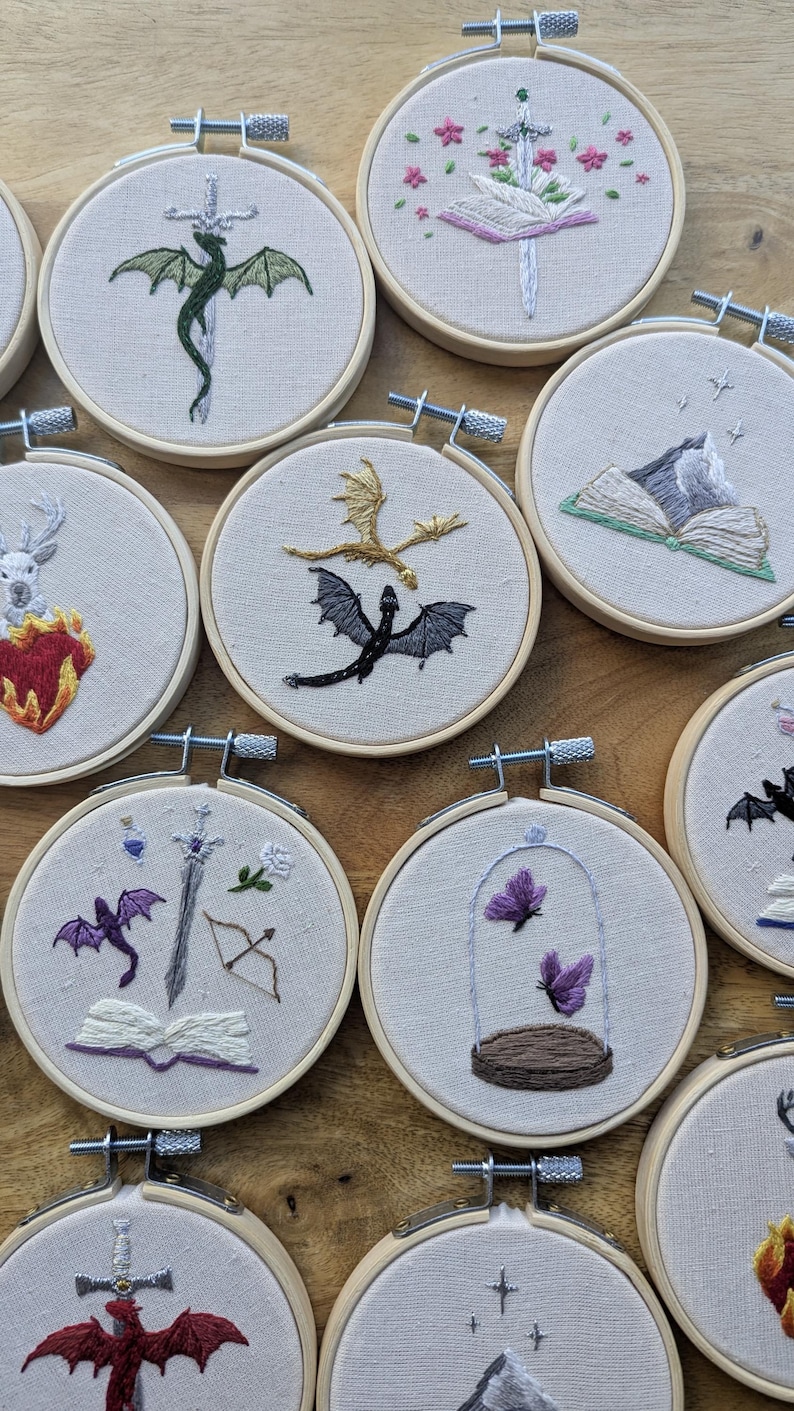 May include: A collection of embroidered hoops featuring fantasy-themed designs. Dragons, swords, books, and butterflies are depicted in colorful thread on a neutral fabric. Each hoop is secured with a metal tightening mechanism.