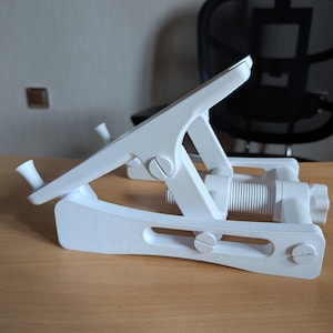 May include: A white, adjustable tablet stand with a clamp design. The stand features a screw mechanism for height adjustment and is made of plastic. It is designed to hold a tablet device and is shown on a wooden desk.