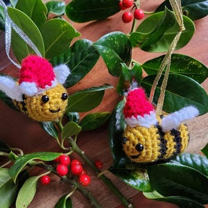 May include: Two crocheted bee ornaments with red and white Santa hats hanging from a holly branch with red berries.
