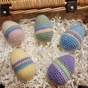 May include: Five crocheted Easter eggs in a wicker basket. The eggs are in pastel colours with stripes of pink, blue, green, and yellow.