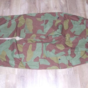 May include: Camouflage pants with suspenders. The pants are made of a brown, green, and tan fabric with a camouflage pattern. The pants have two large pockets on the front and two smaller pockets on the back. The suspenders are made of a brown fabric and have a buckle closure.