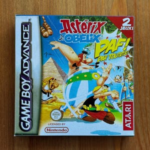 Può includere: Una scatola di cartucce per videogiochi Game Boy Advance con i personaggi di Asterix e Obelix. La grafica della scatola è colorata, con il titolo in rosso e giallo. Il gioco è concesso in licenza da Nintendo e pubblicato da Atari.
