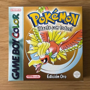 Könnte beinhalten: Eine goldene Game Boy Color-Spielkassette mit einem Bild eines roten, gelben und grünen Vogels mit dem Text "Pokémon ¡Hazte con todos! Edición Oro" auf der Vorderseite.