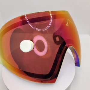 Op de afbeelding: Een rode, iriserende paintball-maskerlens met een witte en roze ring rond de lens.