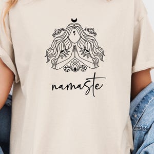 以下が含まれることがあります： ヨガのポーズをとった女性の白黒のイラストが描かれたベージュのTシャツ。イラストの下には「namaste」の文字が書かれている。