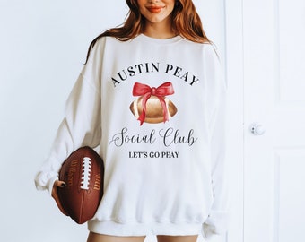 Może przedstawiać: Biała bluza z napisem "Austin Peay Social Club Let's Go Peay" i akwarelową ilustracją piłki nożnej z czerwoną kokardką.