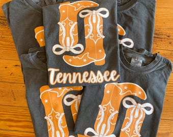 May include: Several charcoal gray t-shirts with a design featuring orange cowboy boots and the word "Tennessee" in white script. The boots have white accents and star details. The shirts are laid out on a wooden surface.