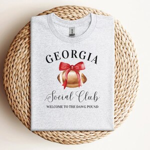 Puede incluir: Camiseta gris con el texto "GEORGIA Social Club WELCOME TO THE DAWG POUND" y una ilustraci&oacute;n de acuarela de un bal&oacute;n de f&uacute;tbol con un lazo rojo.