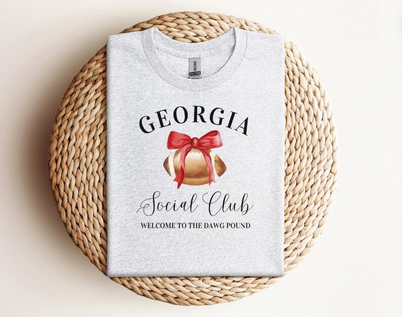 Puede incluir: Camiseta gris con el texto "GEORGIA Social Club WELCOME TO THE DAWG POUND" y una ilustraci&oacute;n de acuarela de un bal&oacute;n de f&uacute;tbol con un lazo rojo.