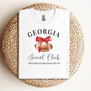 Puede incluir: Camiseta blanca con el texto "GEORGIA Social Club WELCOME TO THE DAWG POUND" y una ilustraci&oacute;n de acuarela de un bal&oacute;n de f&uacute;tbol con un lazo rojo.