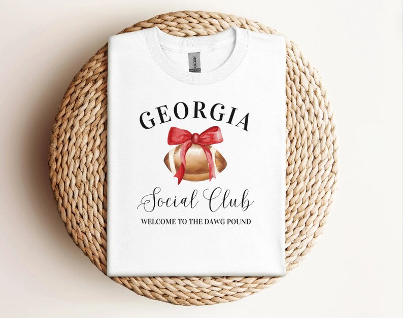 Puede incluir: Camiseta blanca con el texto "GEORGIA Social Club WELCOME TO THE DAWG POUND" y una ilustraci&oacute;n de acuarela de un bal&oacute;n de f&uacute;tbol con un lazo rojo.