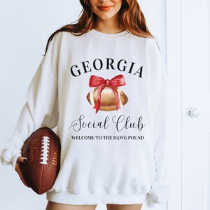 Puede incluir: Sudadera blanca con el texto "GEORGIA Social Club WELCOME TO THE DAWG POUND" y una ilustraci&oacute;n de acuarela de un bal&oacute;n de f&uacute;tbol con un lazo rojo.