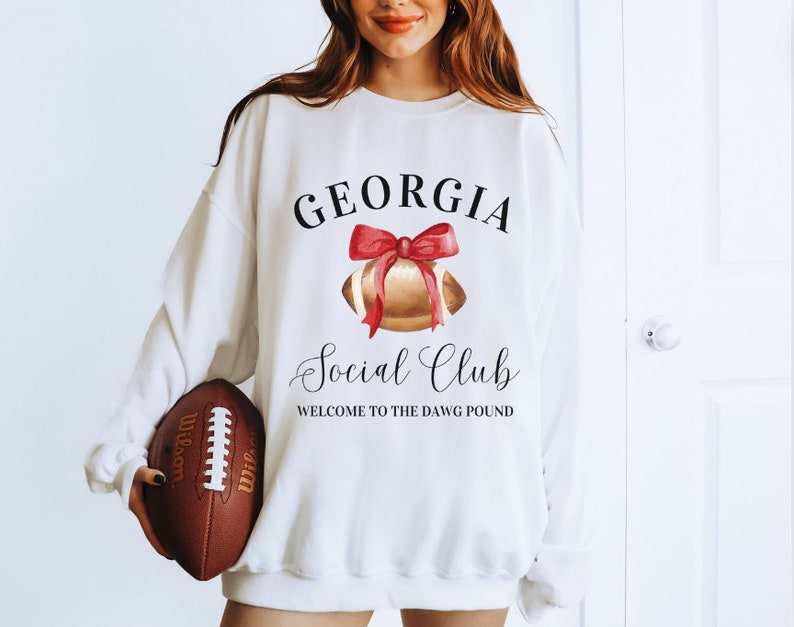 Puede incluir: Sudadera blanca con el texto "GEORGIA Social Club WELCOME TO THE DAWG POUND" y una ilustraci&oacute;n de acuarela de un bal&oacute;n de f&uacute;tbol con un lazo rojo.
