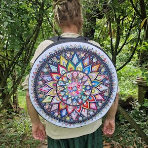 Può includere: Uno zaino nero con un design rotondo di mandala colorato. Il mandala presenta una varietà di colori, tra cui rosso, giallo, blu, verde e viola. Lo zaino è indossato da una persona con i capelli lunghi e intrecciati.
