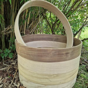 Könnte beinhalten: Ein handgefertigter Holzkorb mit gebogenem Griff. Der Korb besteht aus geschichtetem Holz in verschiedenen Braun- und Beigetönen. Der Korb hat eine zylindrische Form und steht im Freien zwischen grünem Laub.