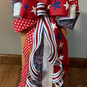May include: A decorative bow featuring red, white, and blue ribbons with various patterns including white stars on red, blue glitter stars, and firework prints. The bow includes long, cascading ribbon tails with striped edges and solid white sections.