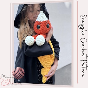 May include: A plush crochet toy shaped like a candy corn with a white pointed top, an orange middle section, and a yellow bottom. The toy features large black safety eyes, embroidered eyebrows, and two white round hands. Text reads Snuggler Crochet Pattern.