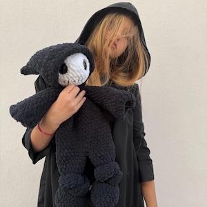 May include: A handmade black crochet doll in the shape of a grim reaper, featuring a white skull face and a hooded cloak. The doll has arms and legs and is held by a person wearing a black hooded cloak.