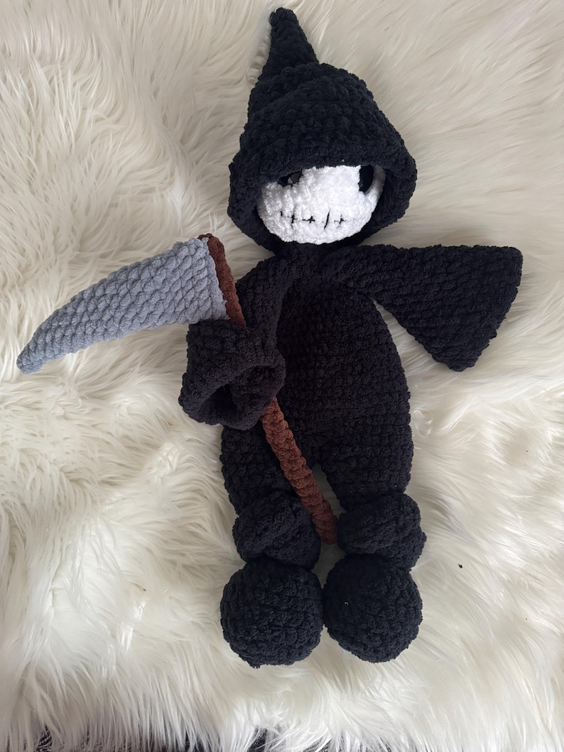 May include: A handmade crochet Grim Reaper doll. The doll is primarily black, with a white skull face and a gray scythe. The scythe has a brown handle. The doll is posed on a white, fluffy surface.