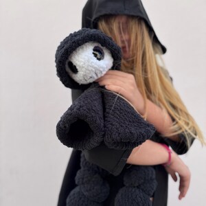May include: A black crocheted doll with a white face and large eyes, wearing a black hooded cloak. The doll has a rounded body and is held against a light background. The doll's features include a black hood and a black body.