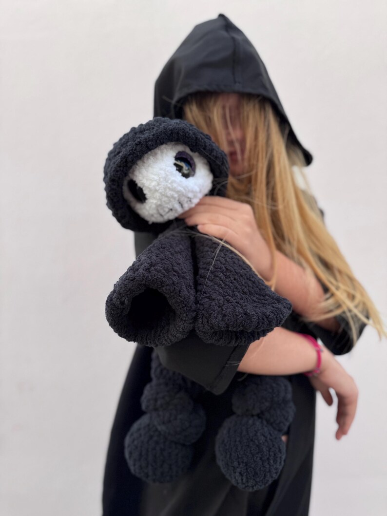 May include: A black crocheted doll with a white face and large eyes, wearing a black hooded cloak. The doll has a rounded body and is held against a light background. The doll's features include a black hood and a black body.
