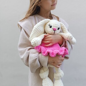 May include: A young girl holds a white crocheted bunny wearing a pink ruffled dress. The bunny has black eyes and a pink nose.