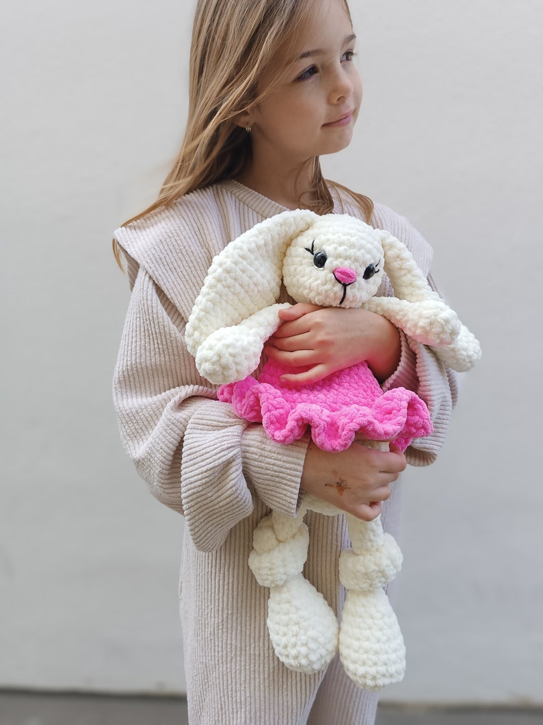May include: A young girl holds a white crocheted bunny wearing a pink ruffled dress. The bunny has black eyes and a pink nose.