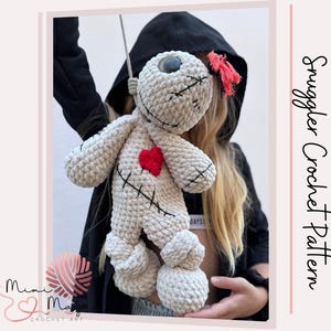 Könnte beinhalten: Eine cremefarbene, gehäkelte Puppe mit einem roten Herz und schwarzen Nahtdetails. Die Puppe hat ein einzelnes Knopfauge und eine kleine rote Quaste. Der Text "Snuggler Crochet Pattern" ist auf der rechten Seite des Bildes zu sehen.