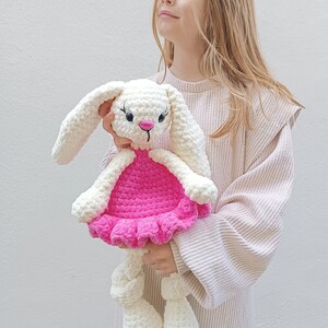 May include: A white crocheted bunny wearing a pink dress. The bunny has black eyes and a pink nose. The bunny is being held by a child.