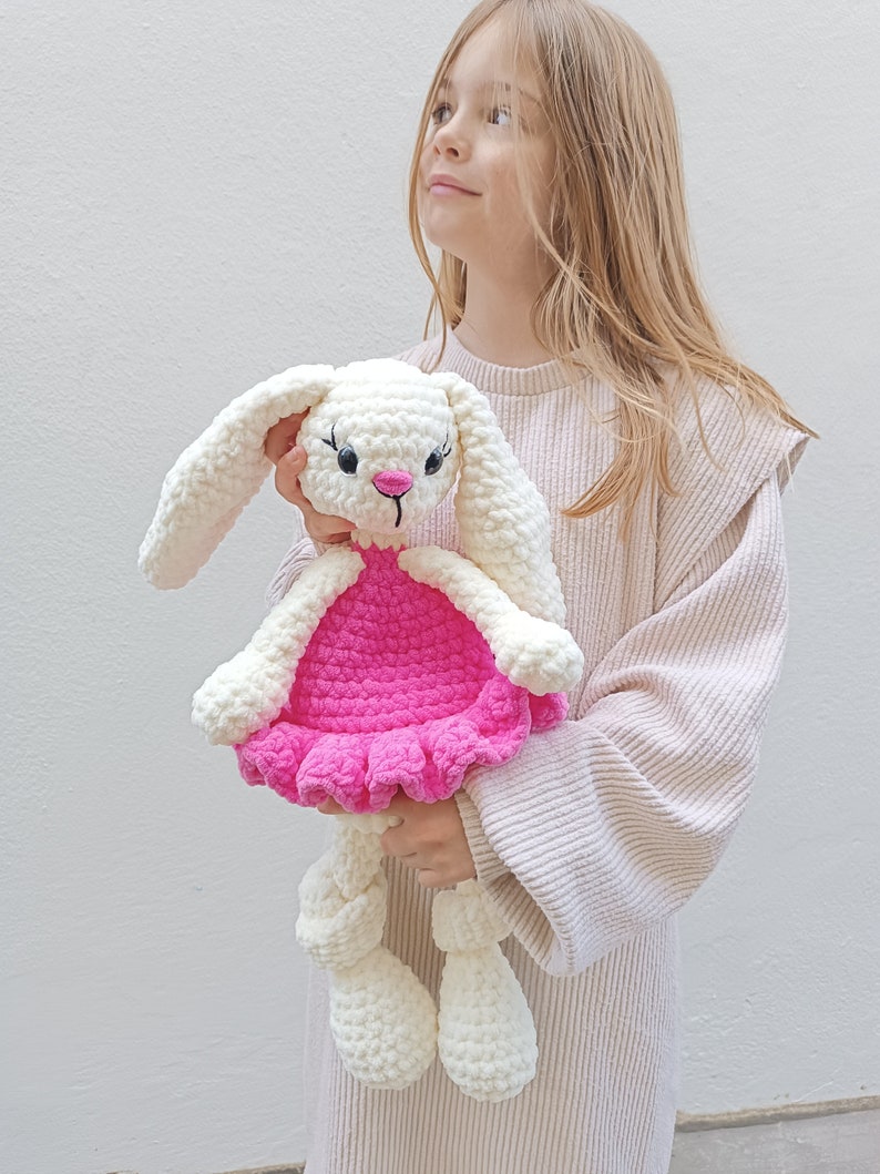 May include: A white crocheted bunny wearing a pink dress. The bunny has black eyes and a pink nose. The bunny is being held by a child.