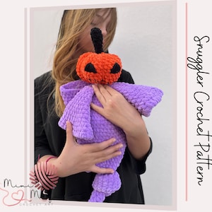 Può includere: Una bambola di zucca all'uncinetto fatta a mano con uno stelo nero e dettagli del viso, tenuta da una persona. La bambola ha una testa arancione vibrante e un corpo viola morbido. Il testo "Snuggler Crochet Pattern" è visibile.