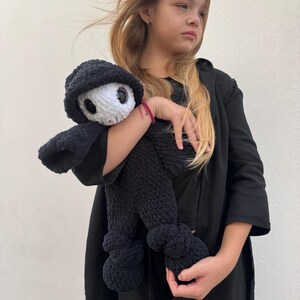 May include: A black crocheted doll with a white face and large eyes, wearing a black hooded outfit. The doll has a rounded body and is being held. The doll's design is reminiscent of a cartoon character.