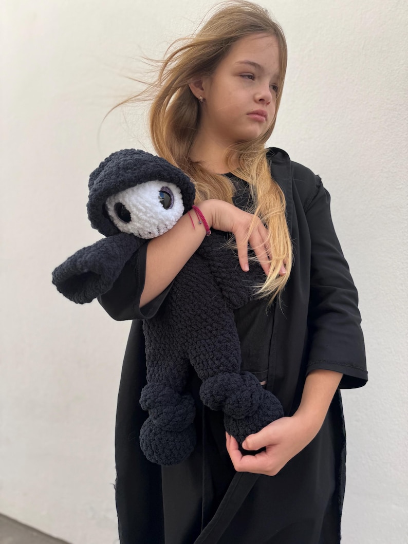 May include: A black crocheted doll with a white face and large eyes, wearing a black hooded outfit. The doll has a rounded body and is being held. The doll's design is reminiscent of a cartoon character.