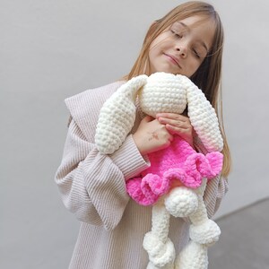 May include: A young girl wearing a beige sweater holds a white crocheted bunny wearing a pink dress. The bunny has large white ears and a fluffy white tail.